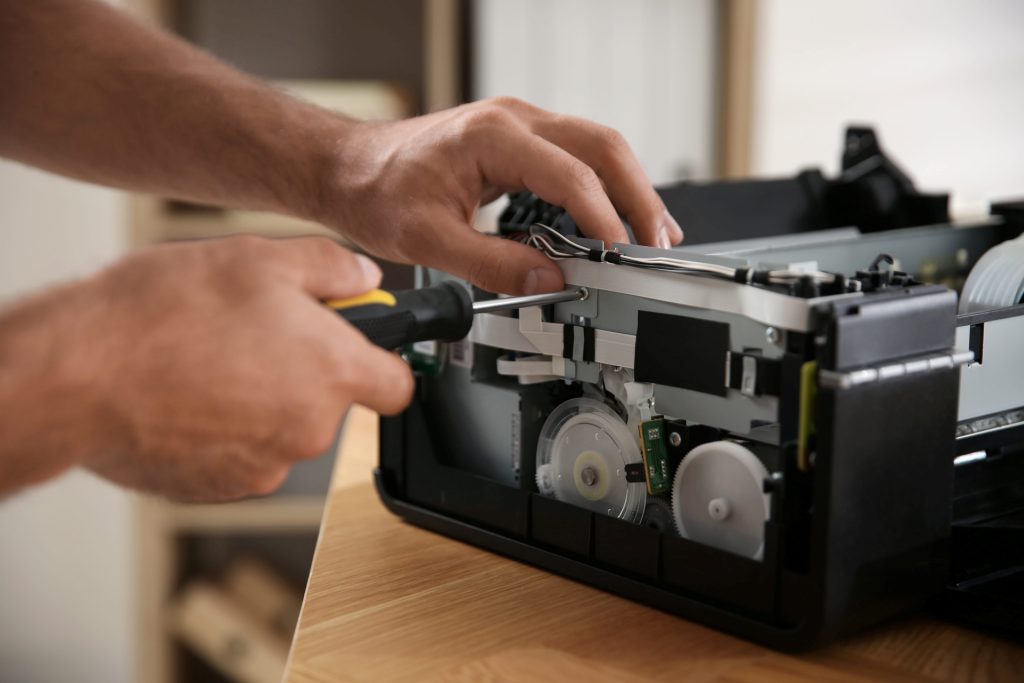 Close-up of hands using a screwdriver to repair the internal components of a printer on a desk.