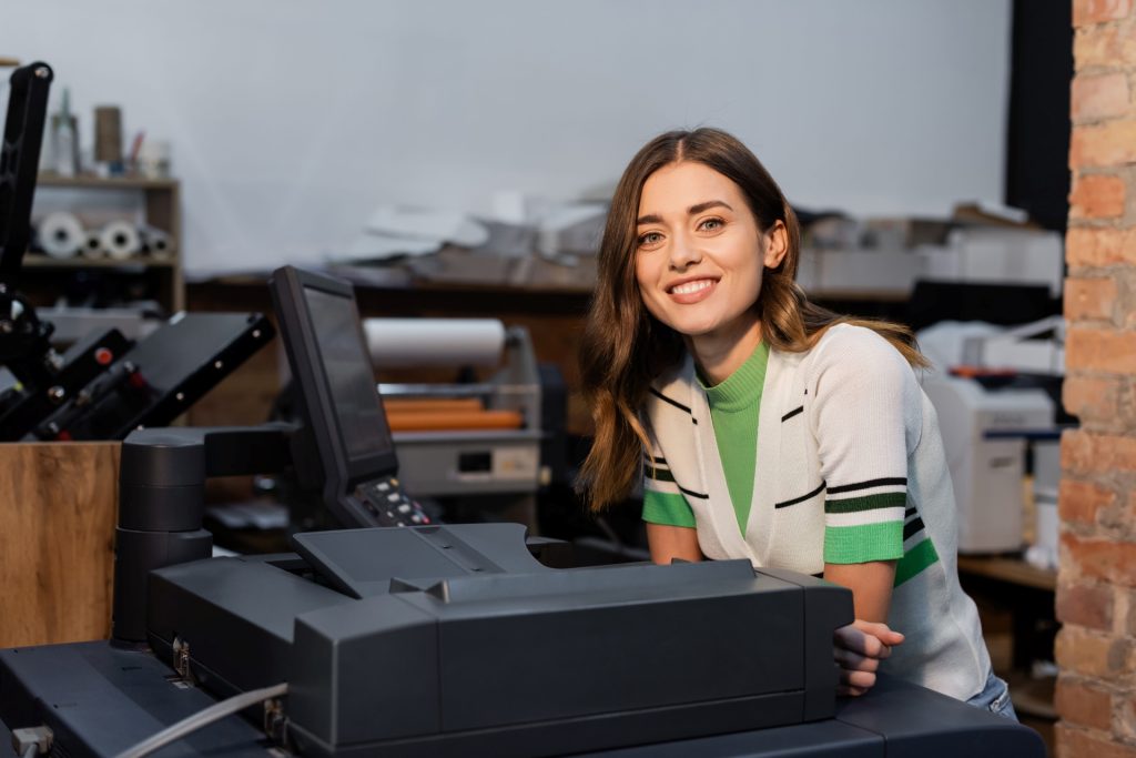 Smiling woman standing beside a large office printer, looking at the camera in a print shop or workspace setting.
