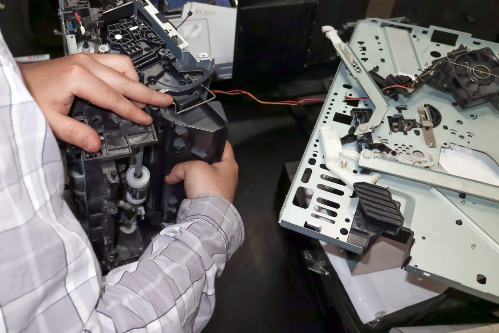 Technician handling and assembling internal printer components, with disassembled parts laid out on a work surface.