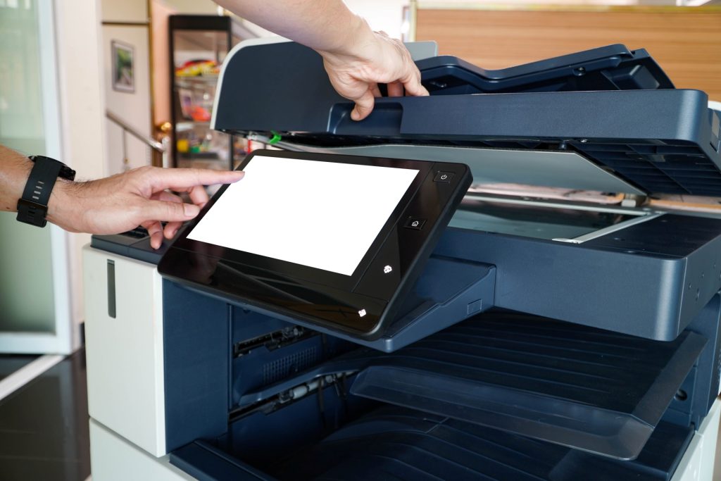 Hands operating a multifunction printer, lifting the scanner lid and using the touchscreen control panel.