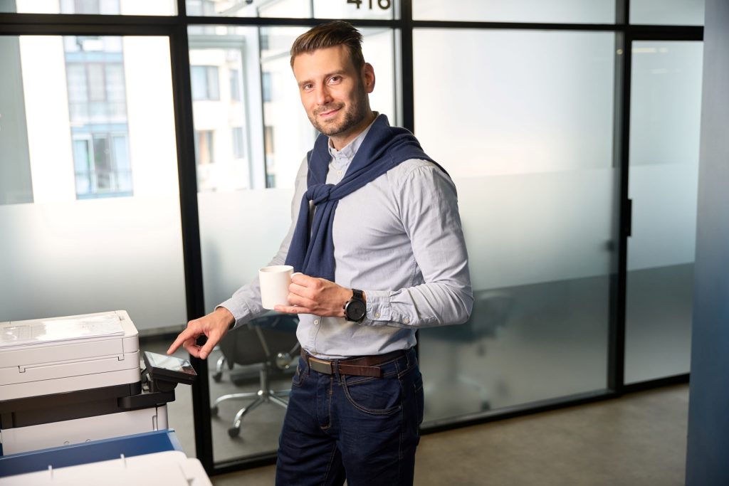 Man holding a coffee cup while using the touchscreen of an office printer in a modern workspace.